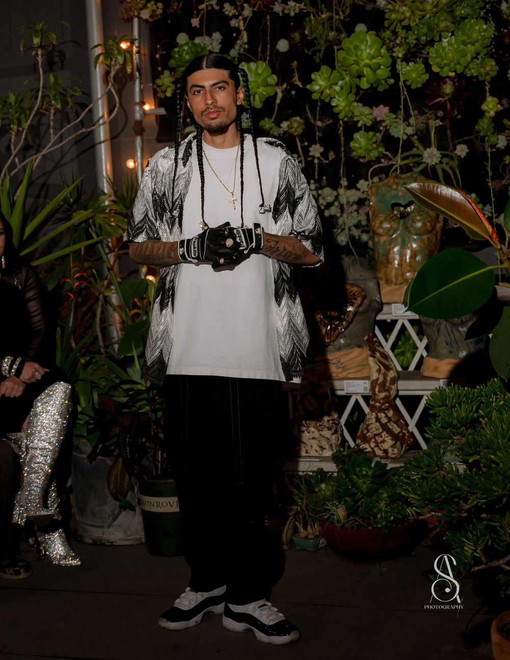 A photographer in a white t-shirt and black patterned kimono stands in a garden setting with plants and decorative lighting.