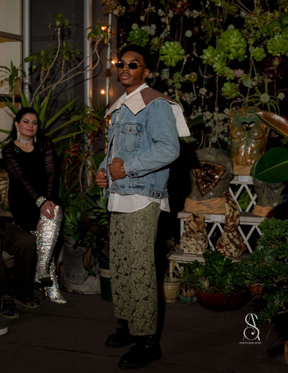 Model wearing denim jacket and floral skirt walks garden runway surrounded by potted plants and string lights.