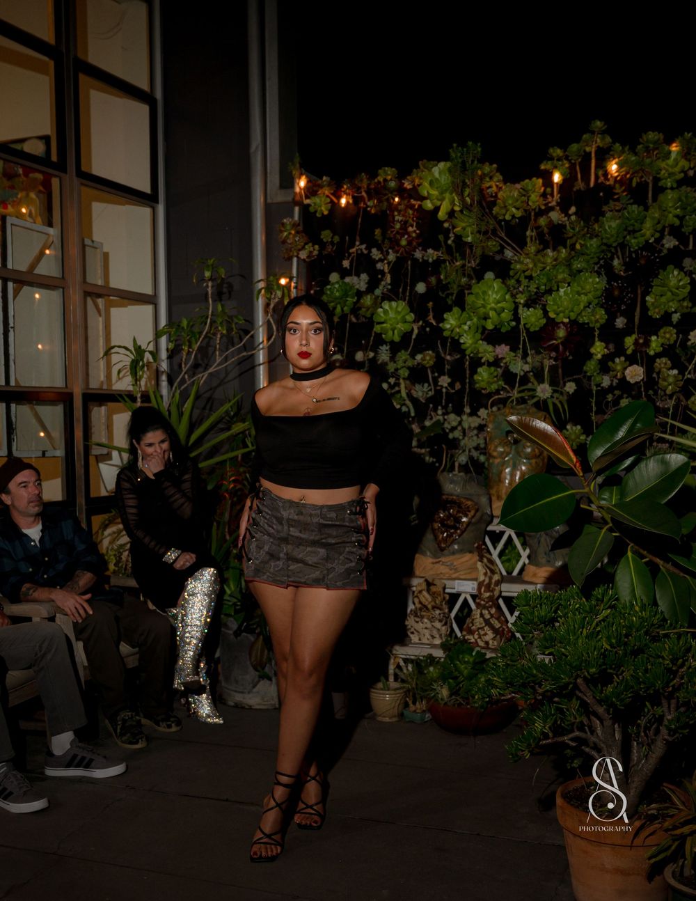 A model walks on a runway surrounded by tropical plants and string lights during an evening fashion show.