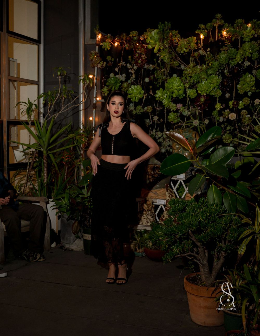 A figure stands in an outdoor garden patio at night surrounded by potted plants and string lights creating a moody ambiance.
