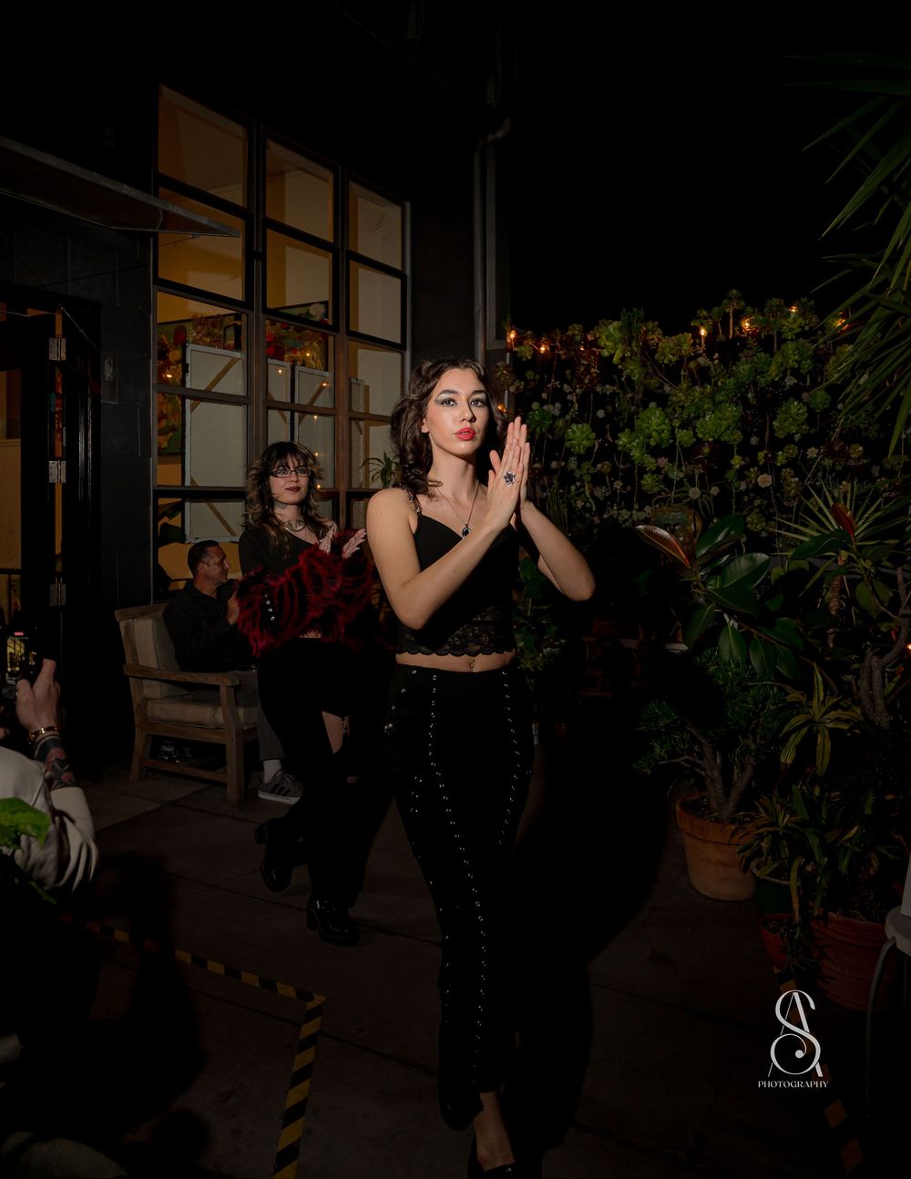 A person in a black outfit strikes a graceful pose during an evening photoshoot on a dimly lit patio with plants.