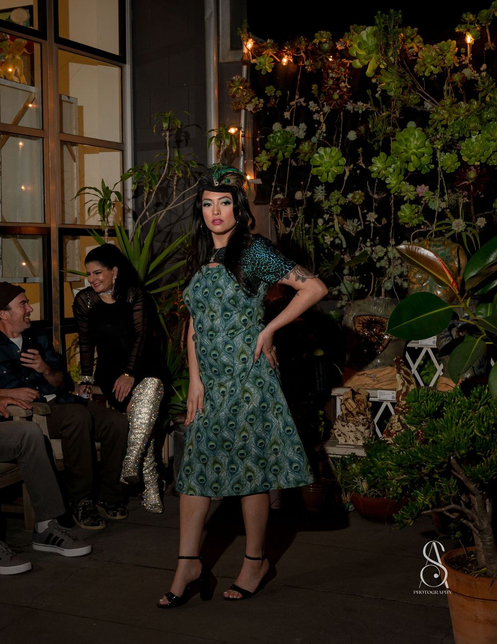 A model poses in a vintage-style green floral dress outside a garden cafe at night with mood lighting and plants.