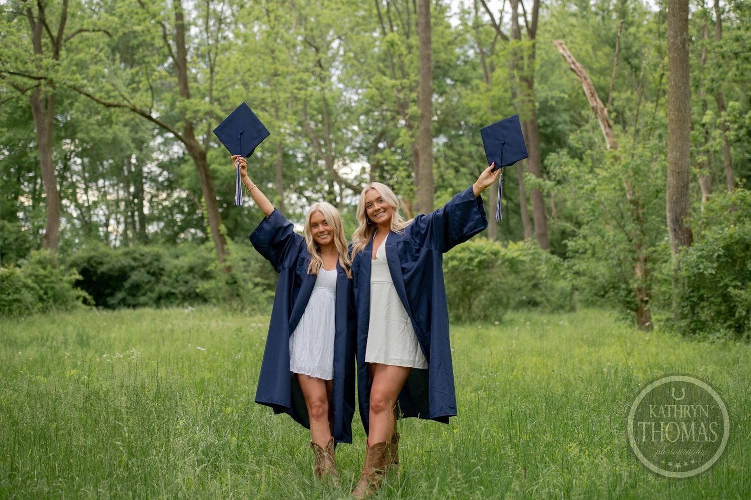 Two graduates in blue caps and gowns celebrate joyfully in a lush green field on graduation day.
