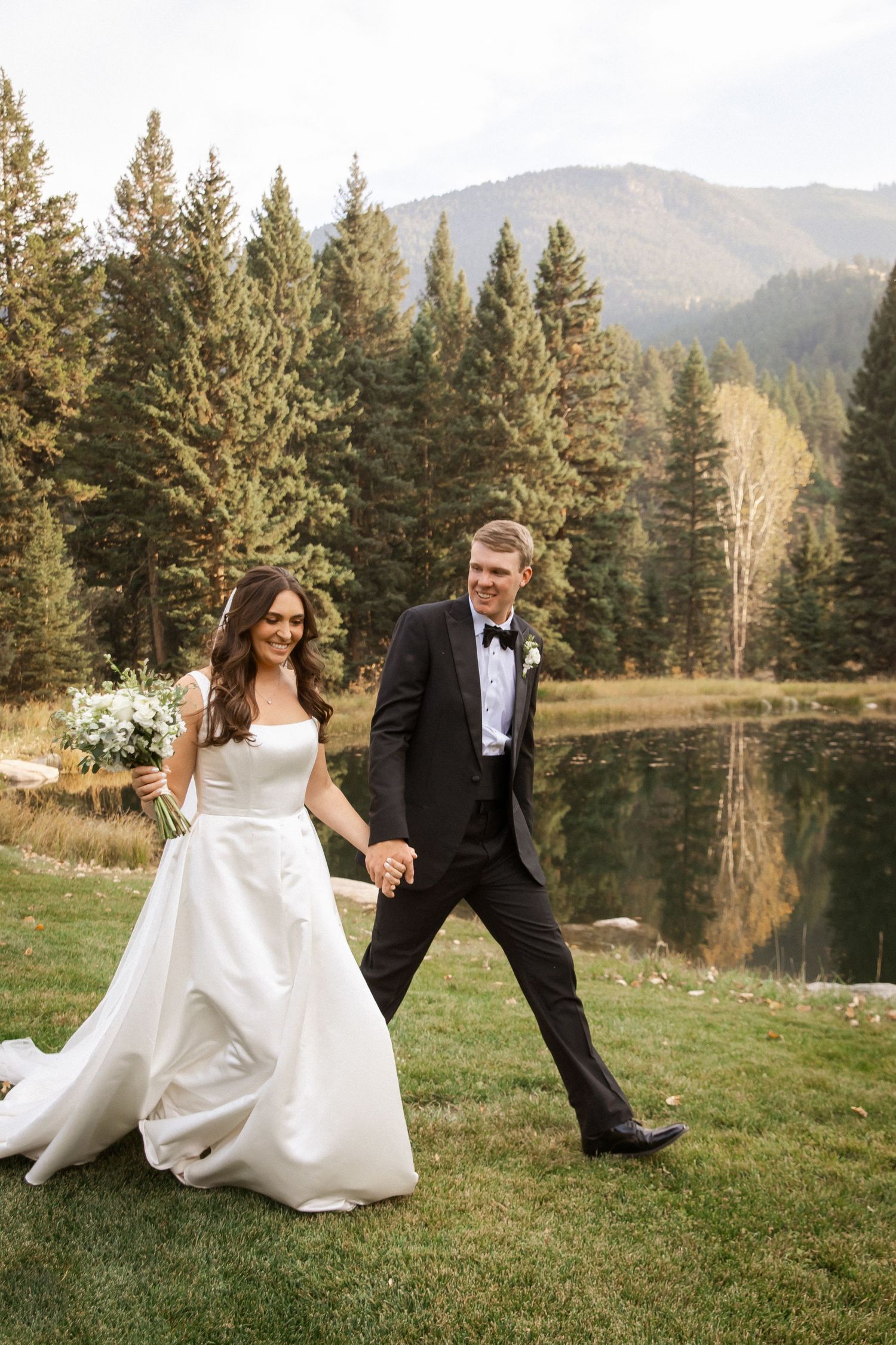 A couple in wedding attire walks together on green grass near a serene lake surrounded by evergreen trees and mountains.
