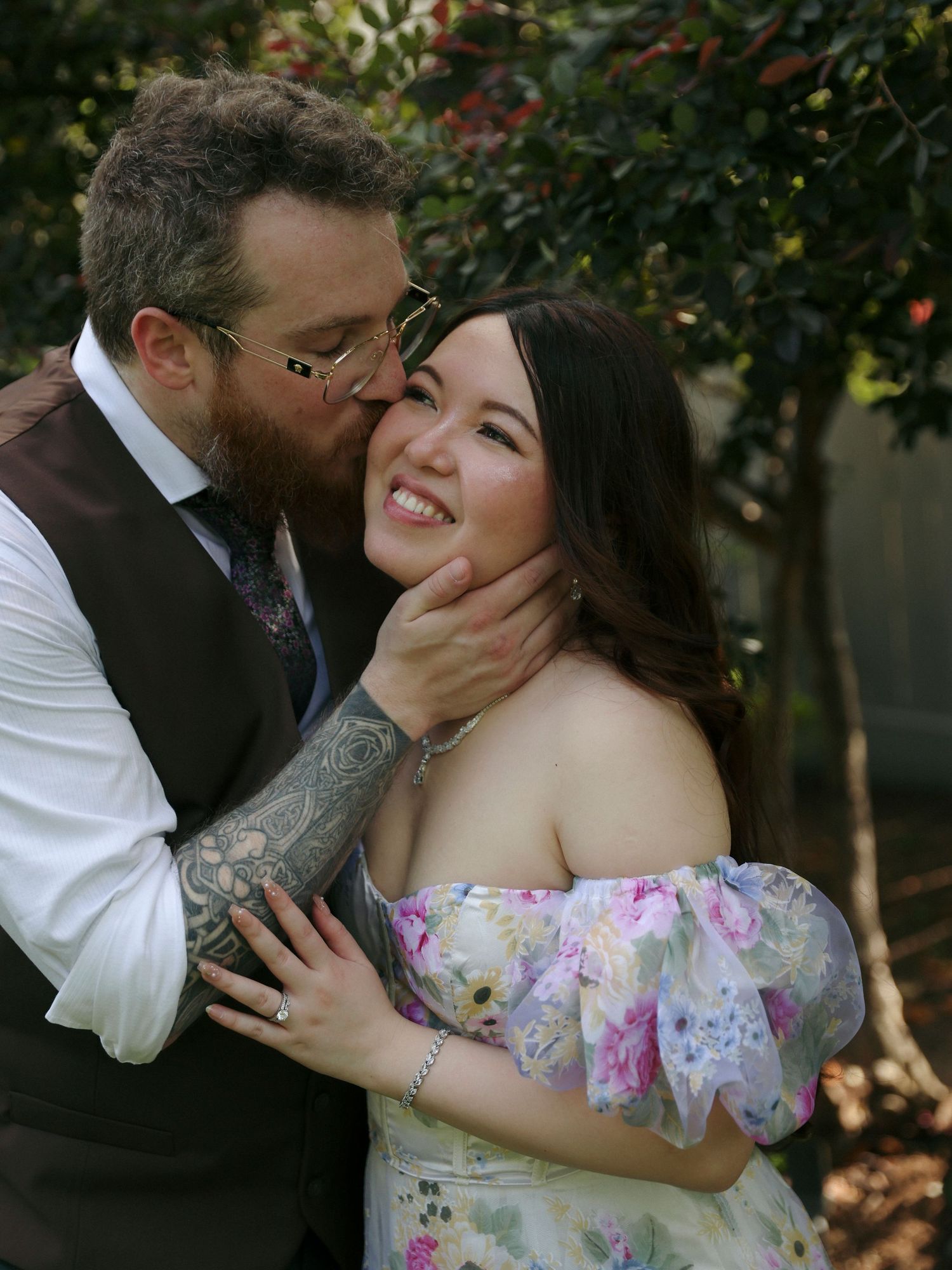 A loving couple share an intimate moment outdoors, with one wearing a floral off-shoulder dress and the other in a vest.