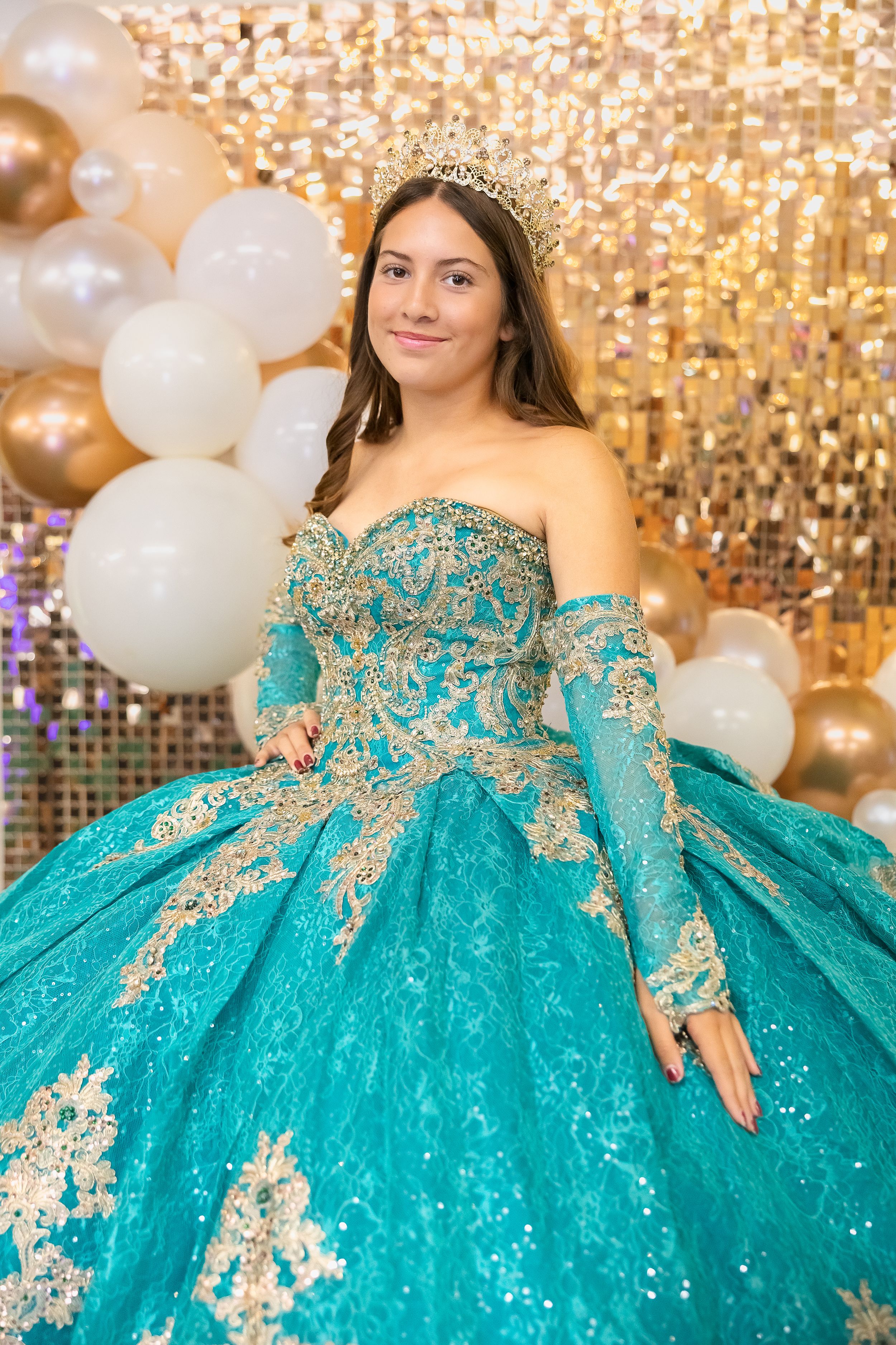 A stunning turquoise and gold quinceañera ball gown sparkles under elegant lighting with white and gold balloon decor.