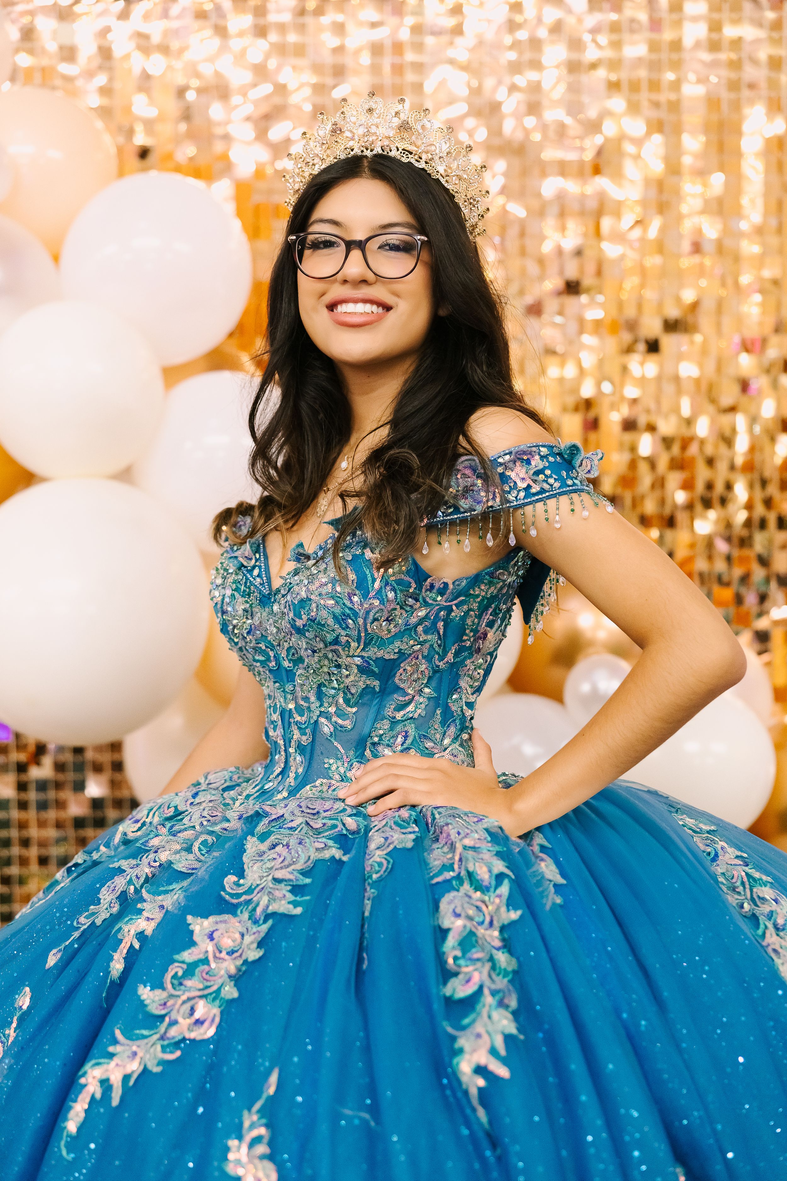 A young woman in glasses wearing a sparkly blue quinceañera ball gown with silver embroidery poses against a gold backdrop.