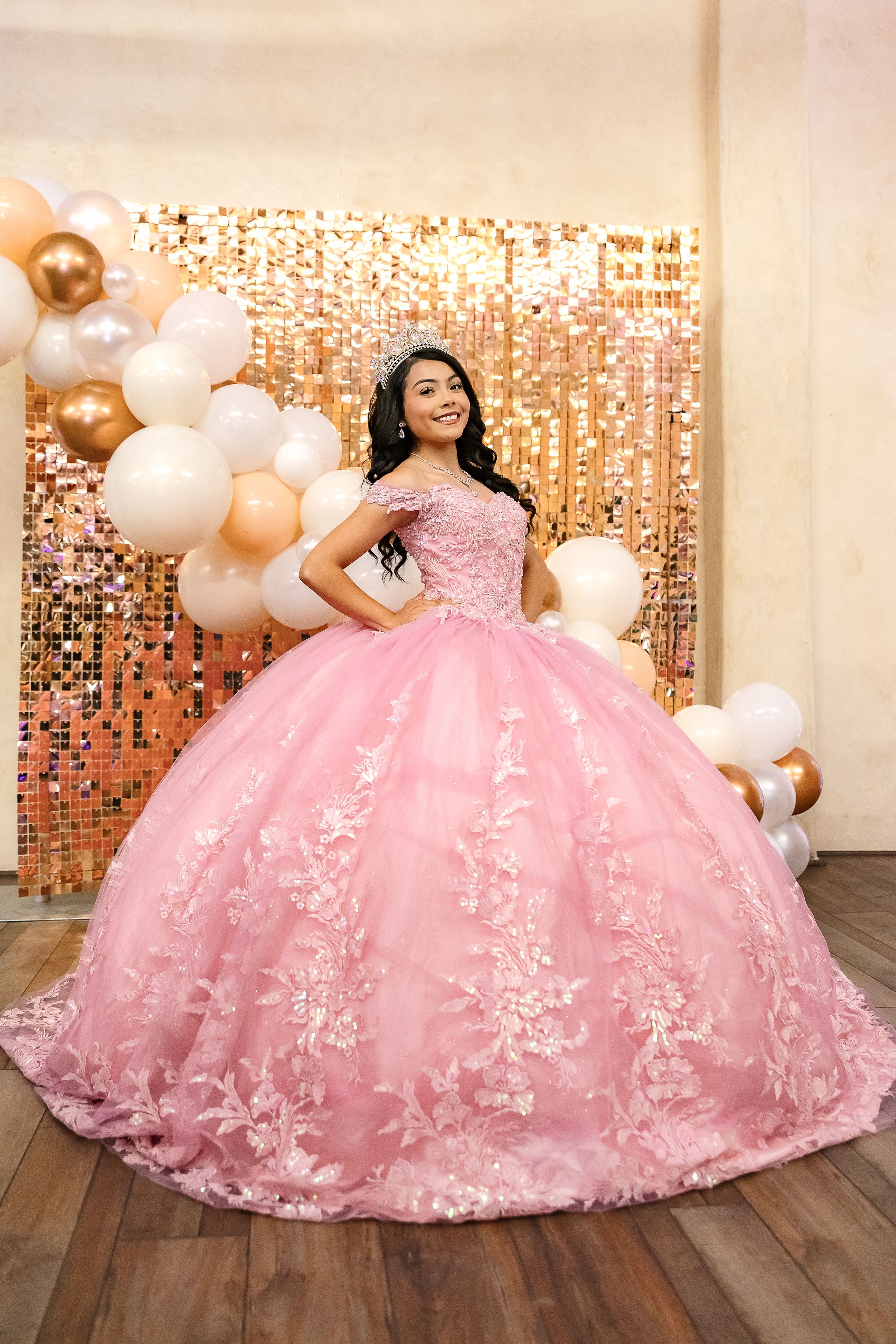 A stunning pink quinceañera ball gown with floral appliques and tulle skirt photographed against a golden backdrop.