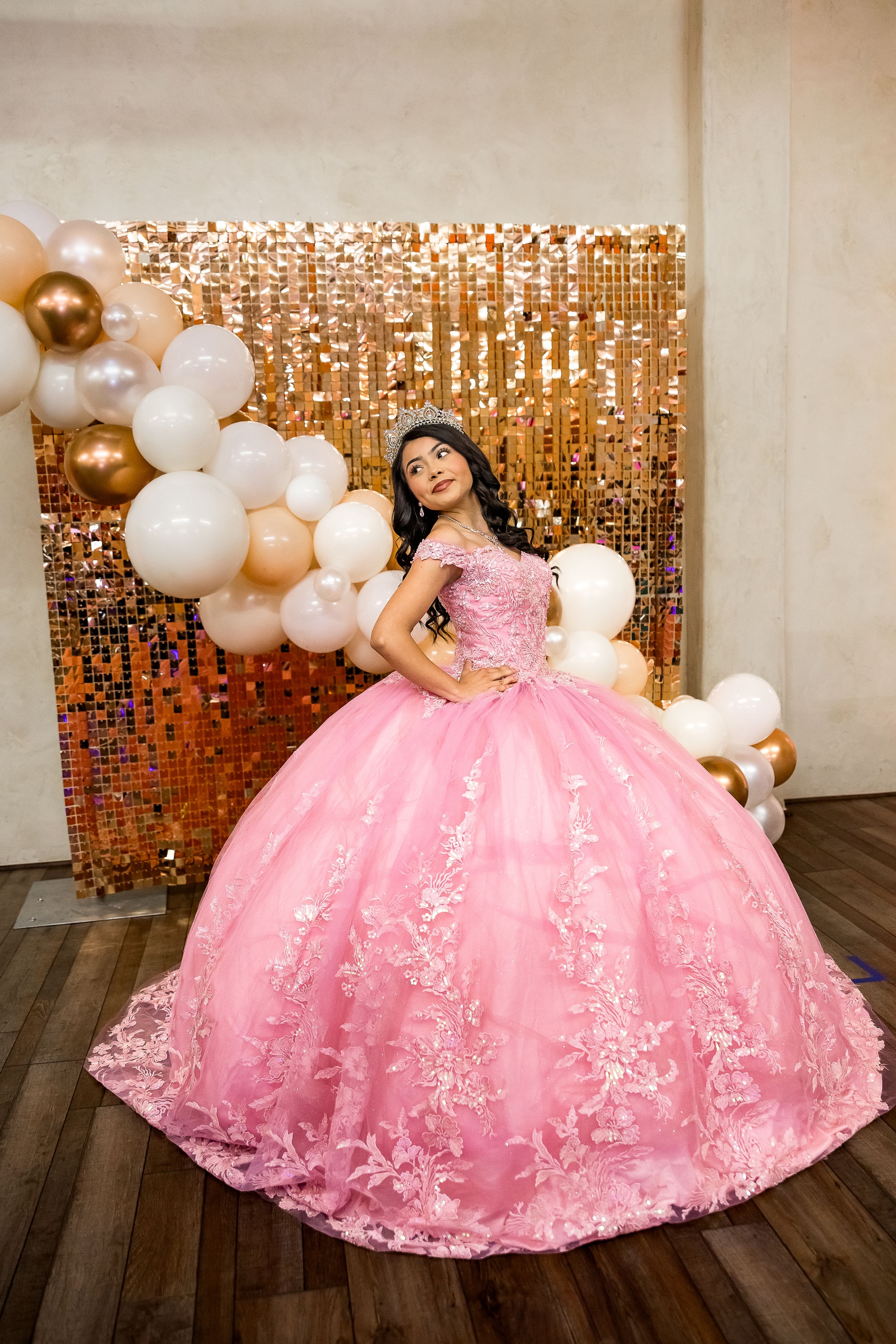 A model poses in a pink ball gown with floral embellishments against a gold sequin backdrop with white and gold balloons.