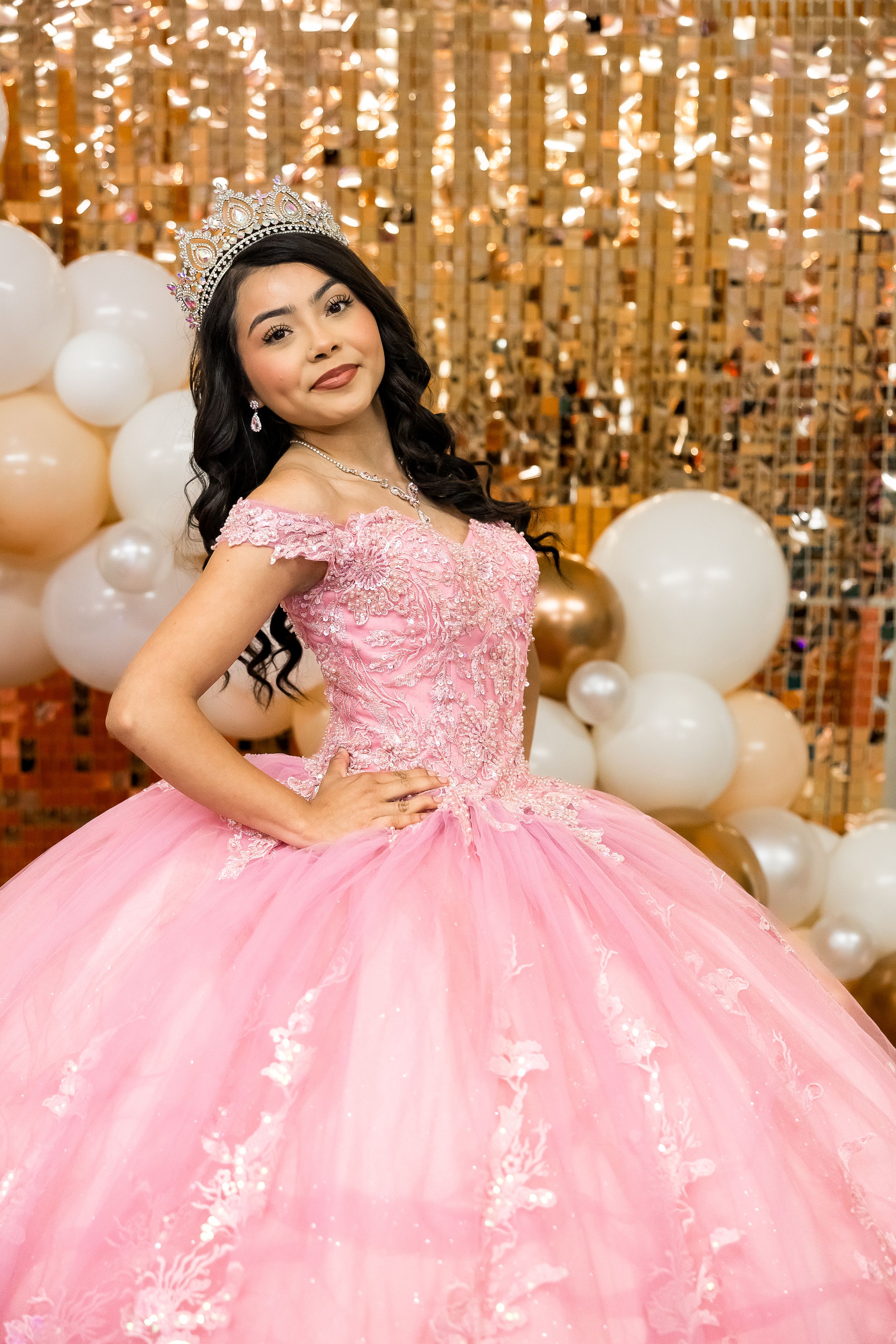 A pink tulle quinceanera dress sparkles against a gold sequin backdrop with white balloons.