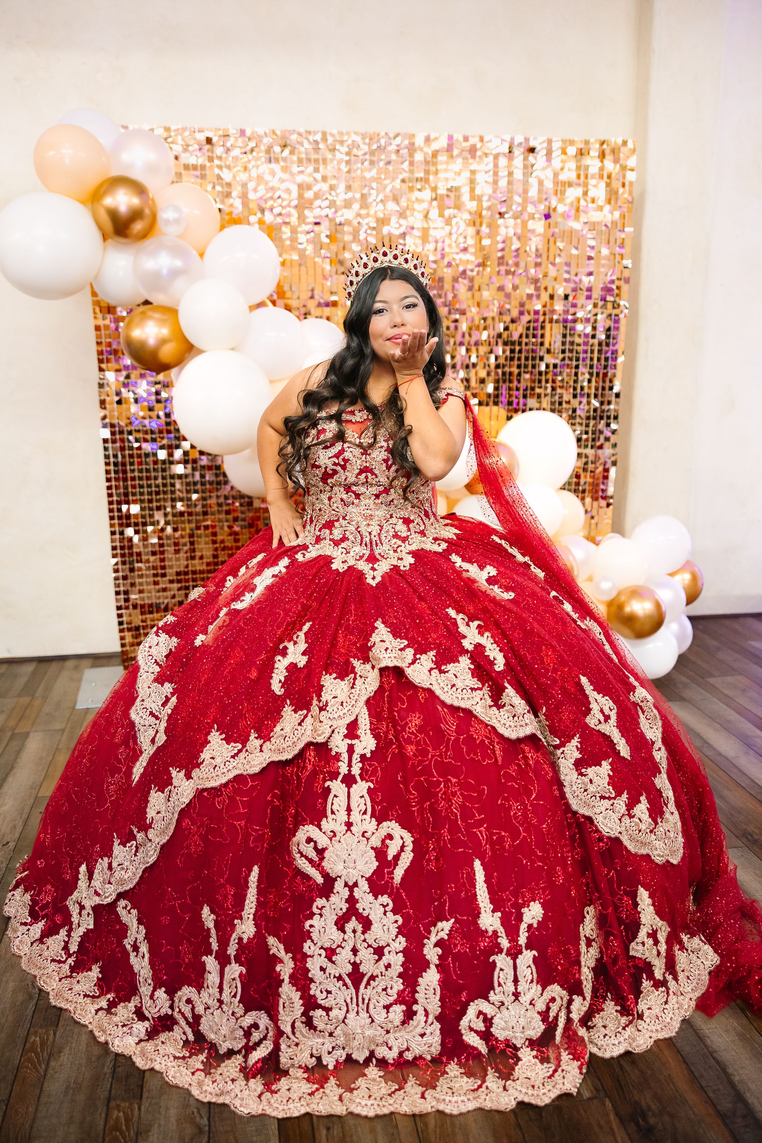 A stunning red quinceanera ball gown with gold lace appliques against a sparkly backdrop with white and gold balloons.