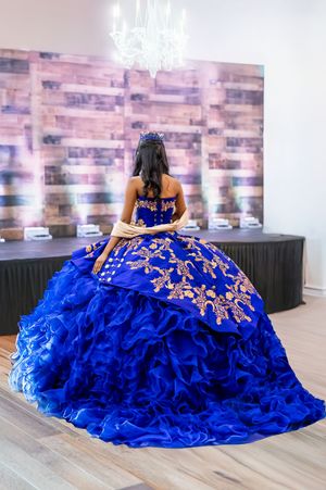 A person in a royal blue quinceañera dress with gold embroidery sits in an elegant room with a chandelier.