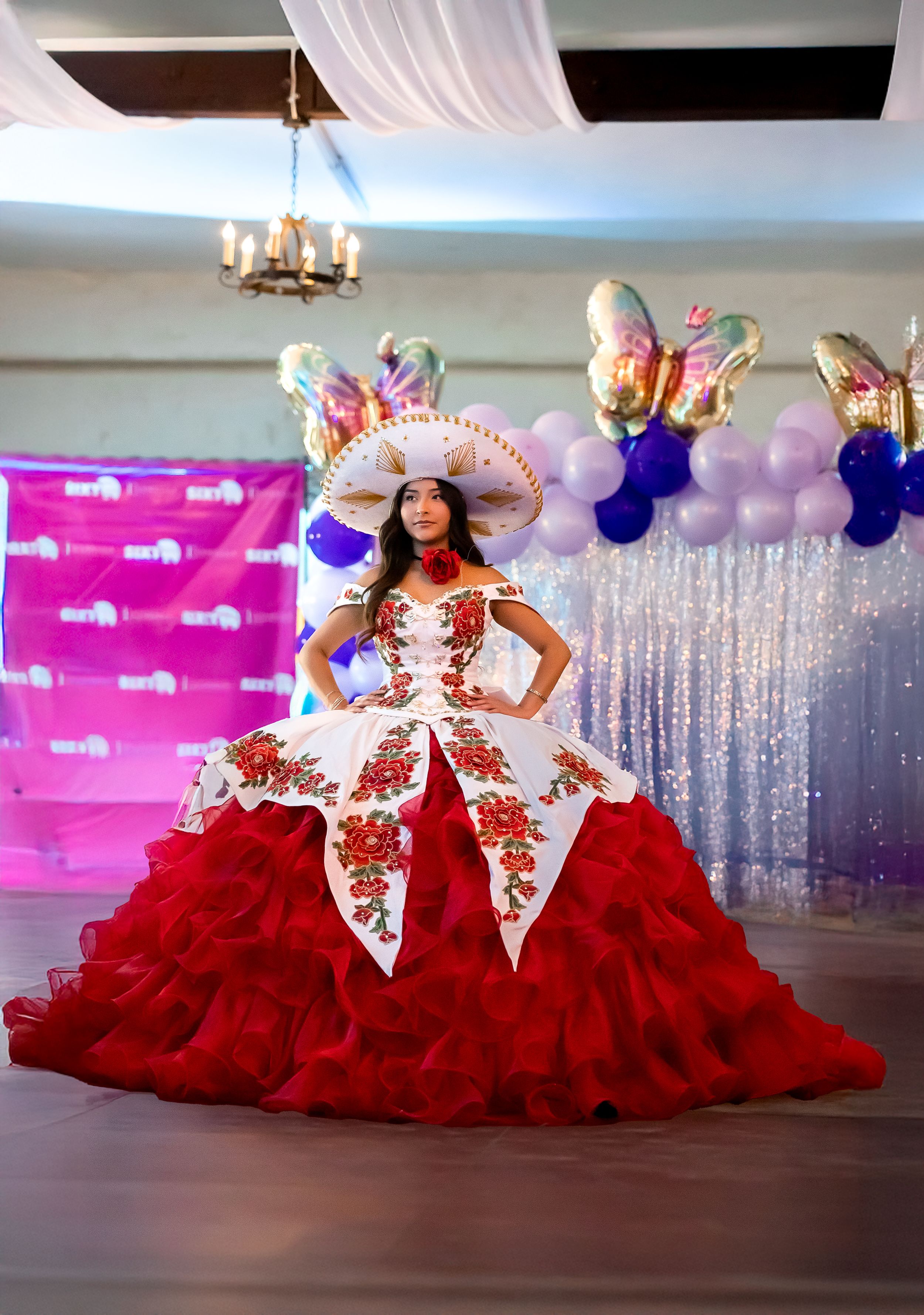 A red and white quinceañera dress with floral details and a matching sombrero at a decorated venue.