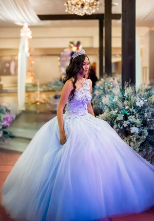 A person in an elegant white and purple quinceañera ball gown poses in front of decorative floral arrangements.
