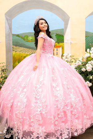An elegant pink quinceanera dress with delicate lace detailing photographed in an outdoor garden setting with arches.