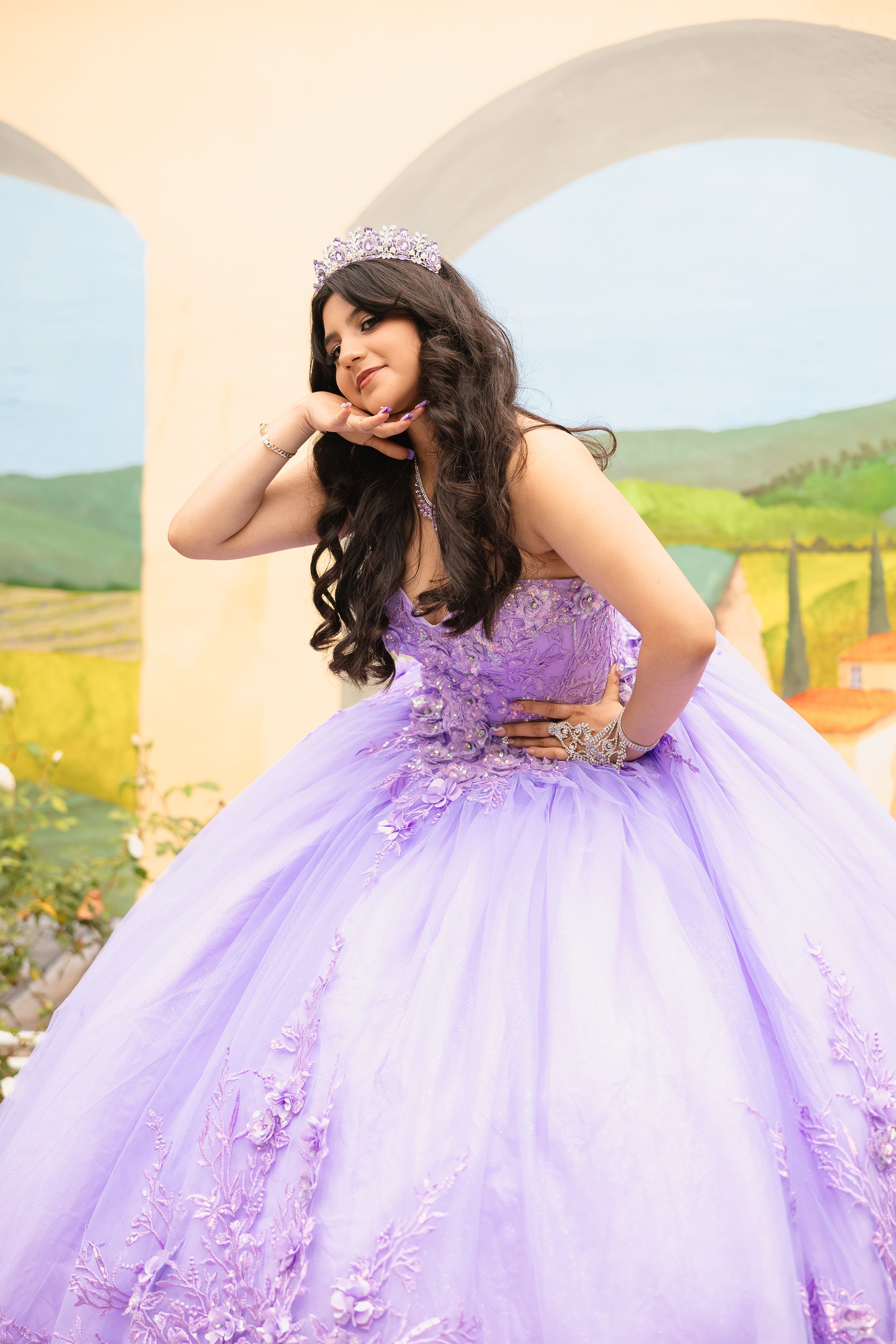 A person in a lavender quinceanera ball gown with embellished details poses outdoors against a pastoral landscape.