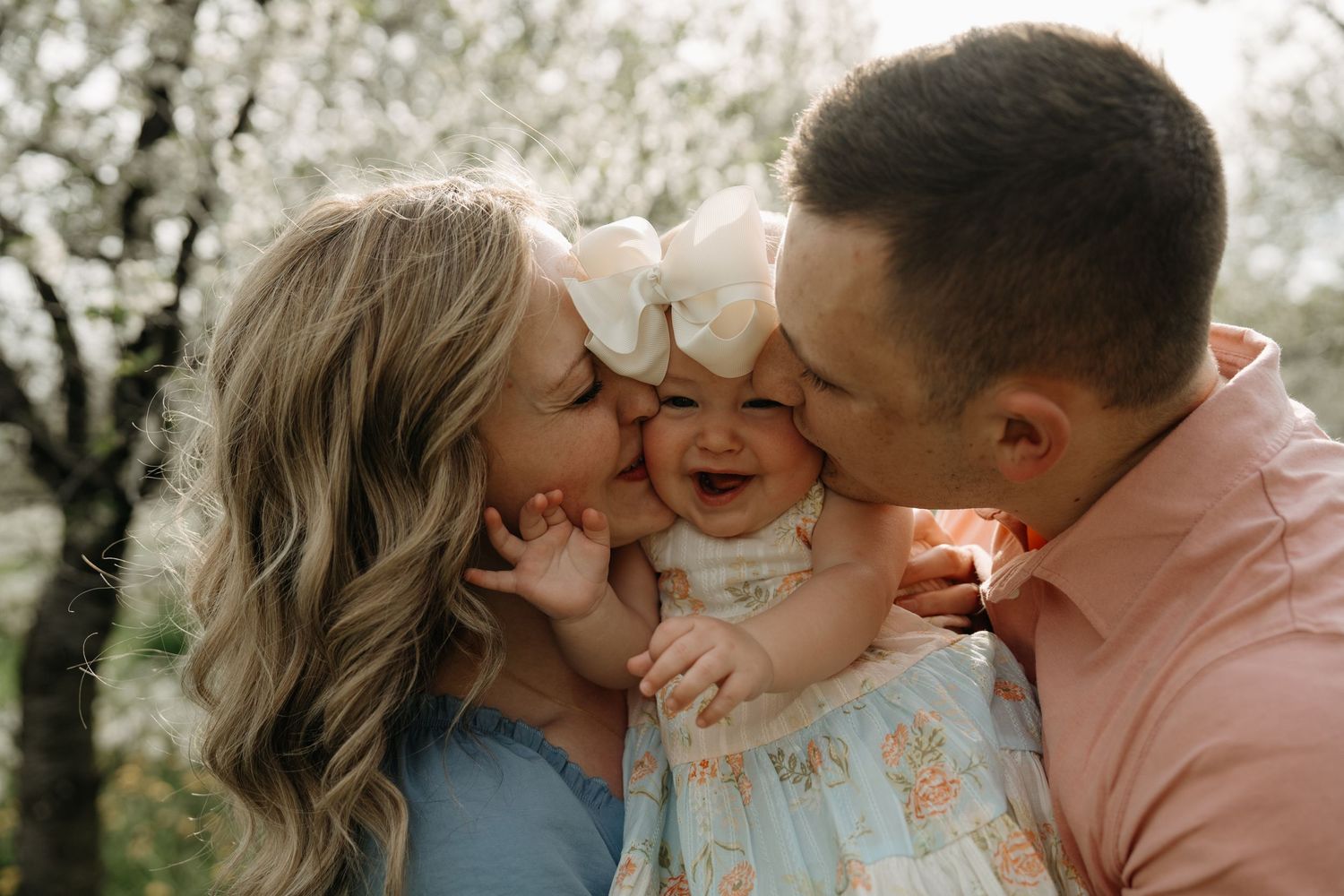 Young parents kiss their giggling child among blooming spring trees during an outdoor family photoshoot.
