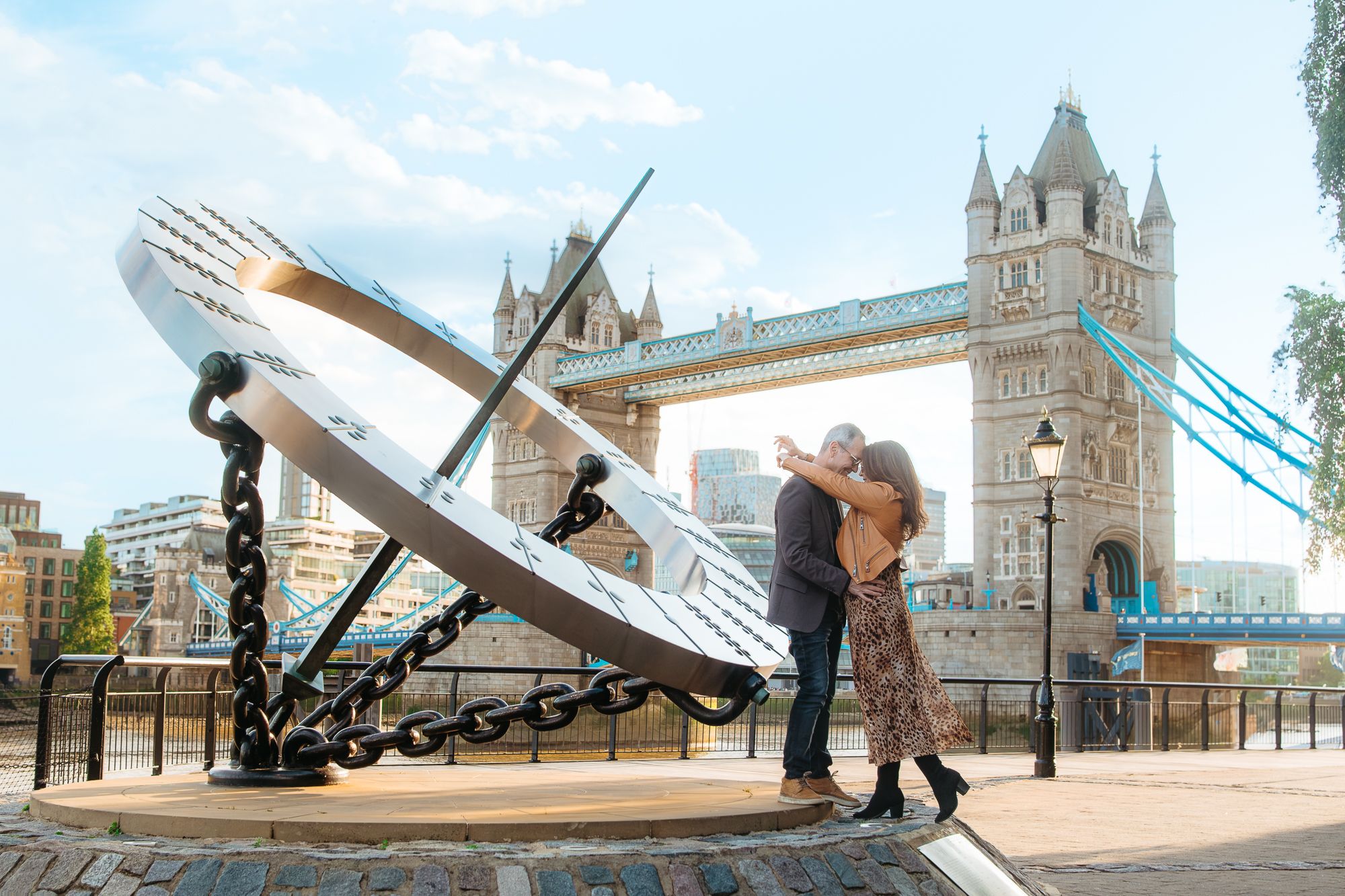 A couple embraces near the iconic Tower Bridge and giant sundial sculpture in London on a sunny day.