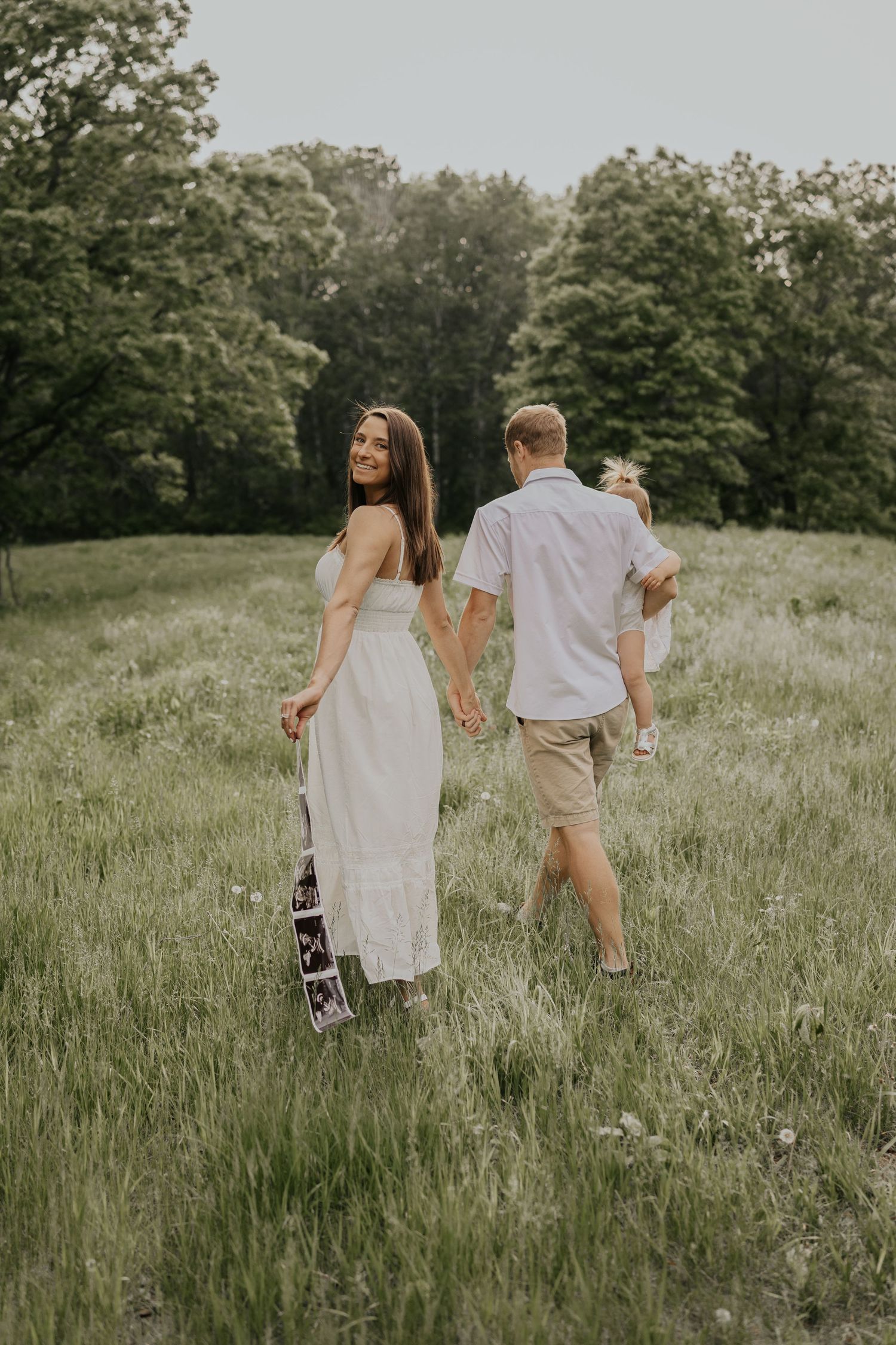 Couple walking hand in hand through a sunny meadow with green trees in the background.