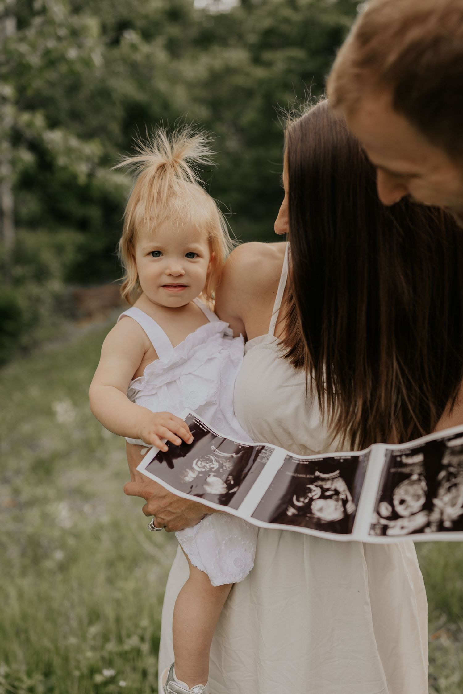 A heartwarming outdoor photo showing ultrasound images being held while embracing a toddler in a white dress.
