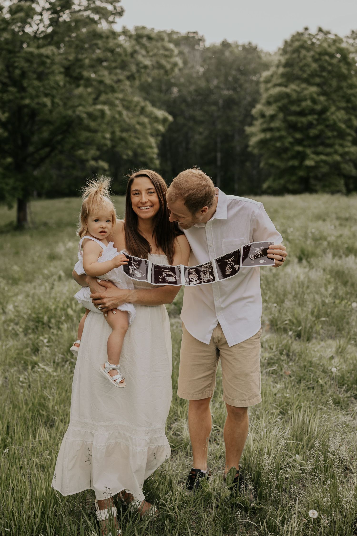 A family stands in a meadow holding a strip of ultrasound photos while wearing casual summer attire.