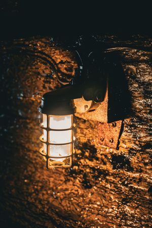 A vintage nautical-style lantern illuminates a rustic stone wall with warm orange light in the darkness.