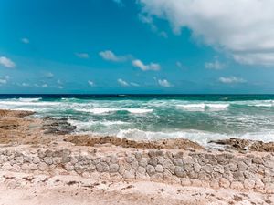 Turquoise waves crash against a stone seawall on a tropical beach under bright blue skies.