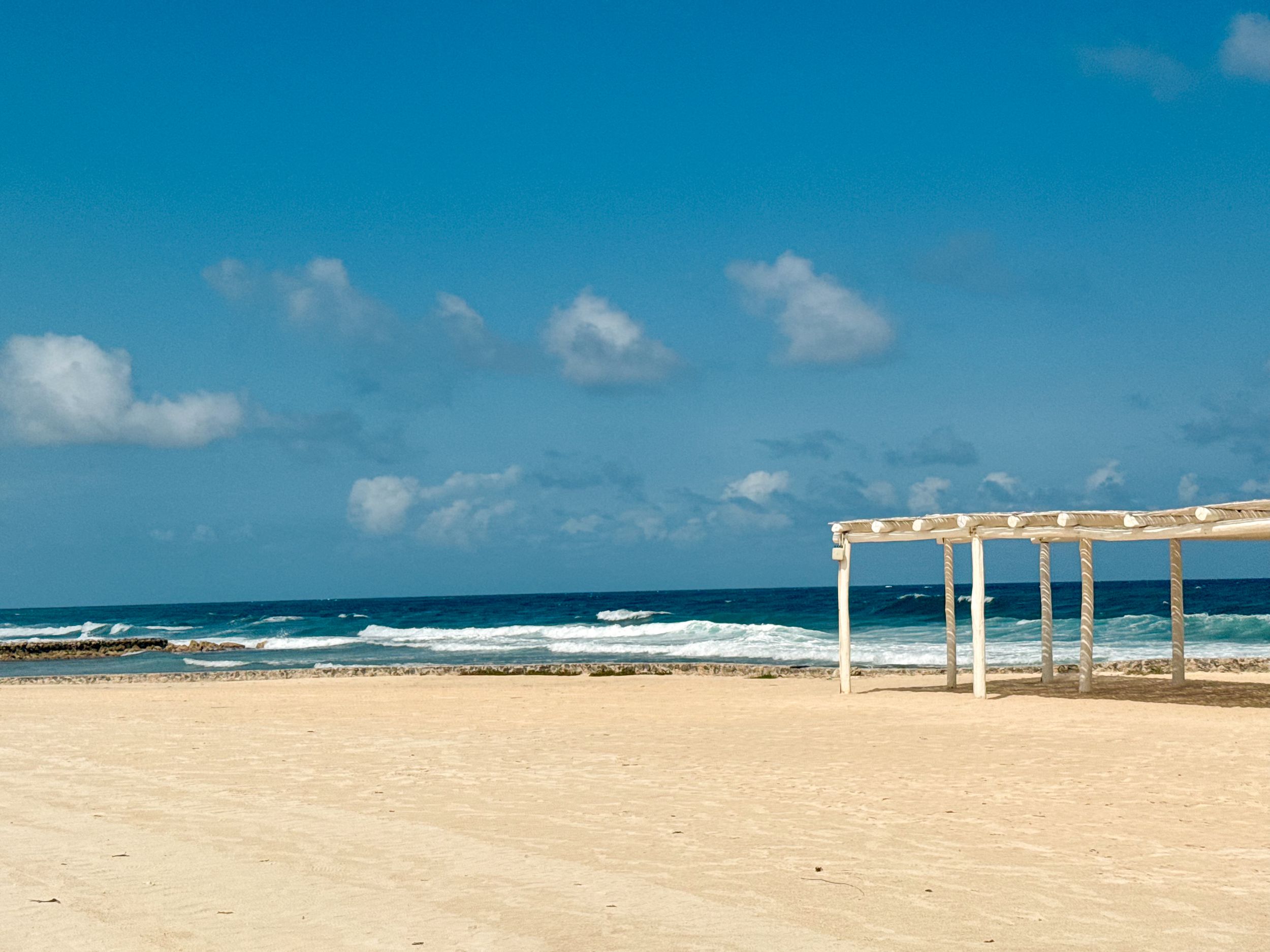 White pergola structure on a pristine sandy beach with blue ocean waves and cloudy sky.