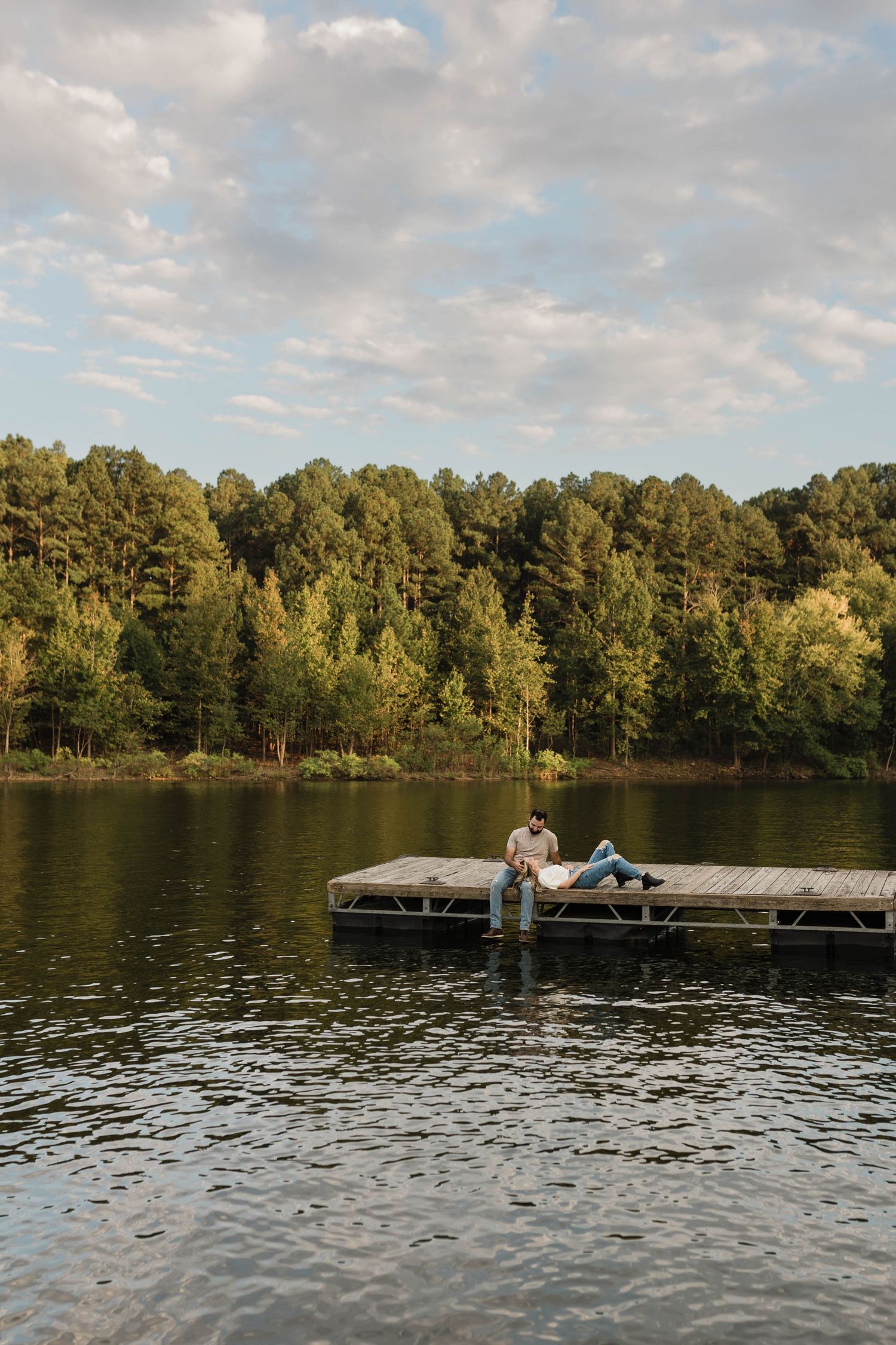 Two people relax on a wooden dock by a peaceful lake surrounded by green trees on a cloudy afternoon.