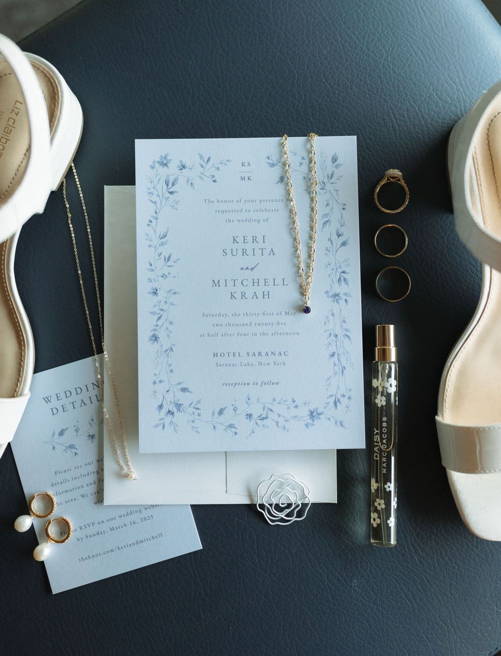 Wedding invitation flatlay with ivory stationery, golden rings, white heels, and delicate floral designs on a dark background.