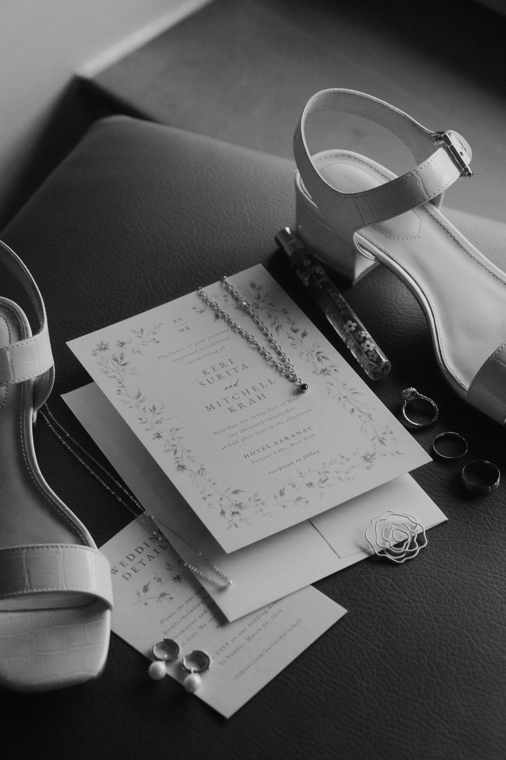 Black and white photo of elegant wedding stationery with ornate details and white sandals on dark leather surface.