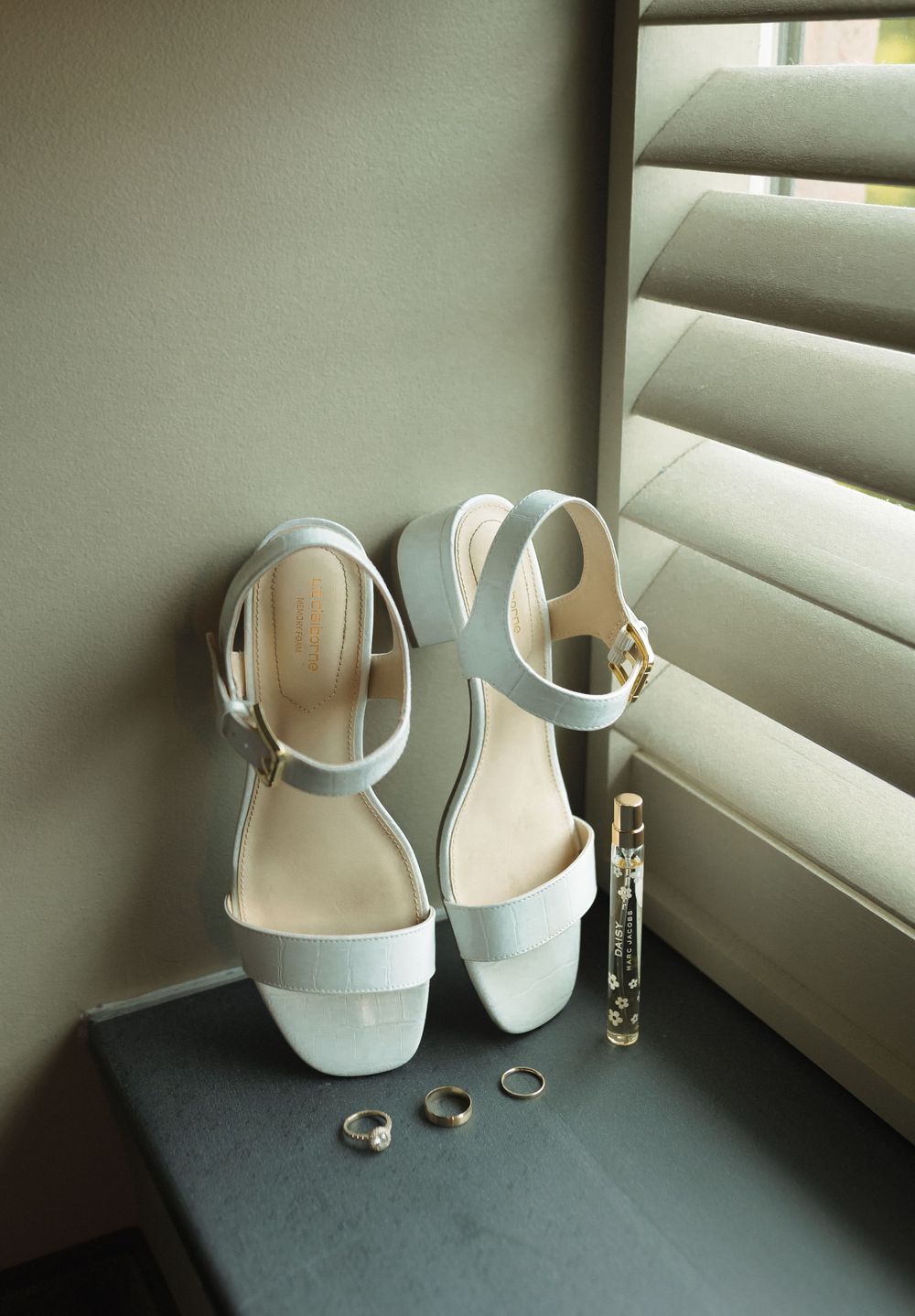 White bridal shoes, perfume bottle, and wedding rings arranged on dark surface next to venetian blinds in natural light.
