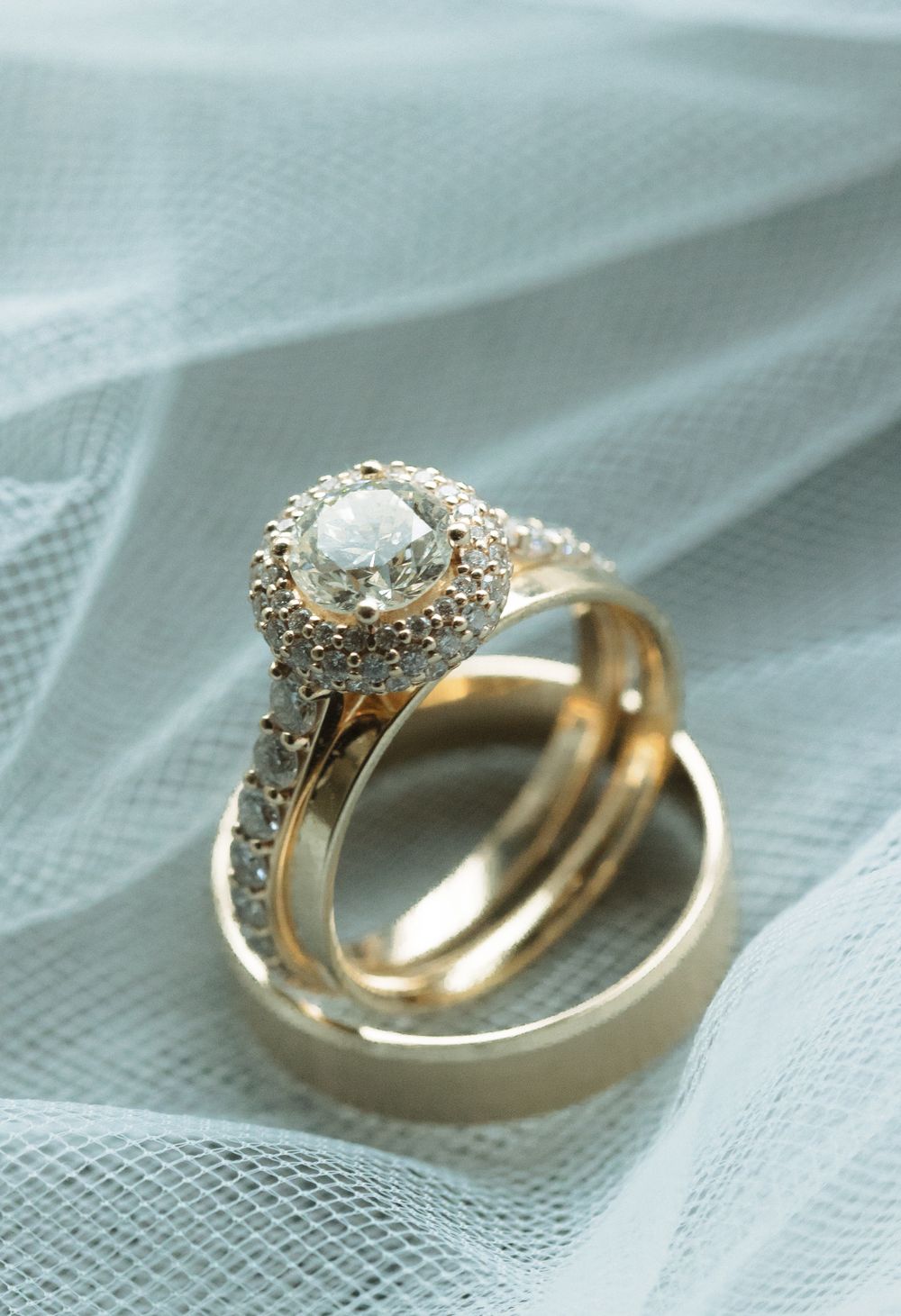 A stunning diamond engagement ring and gold wedding band rest on delicate light blue tulle fabric.