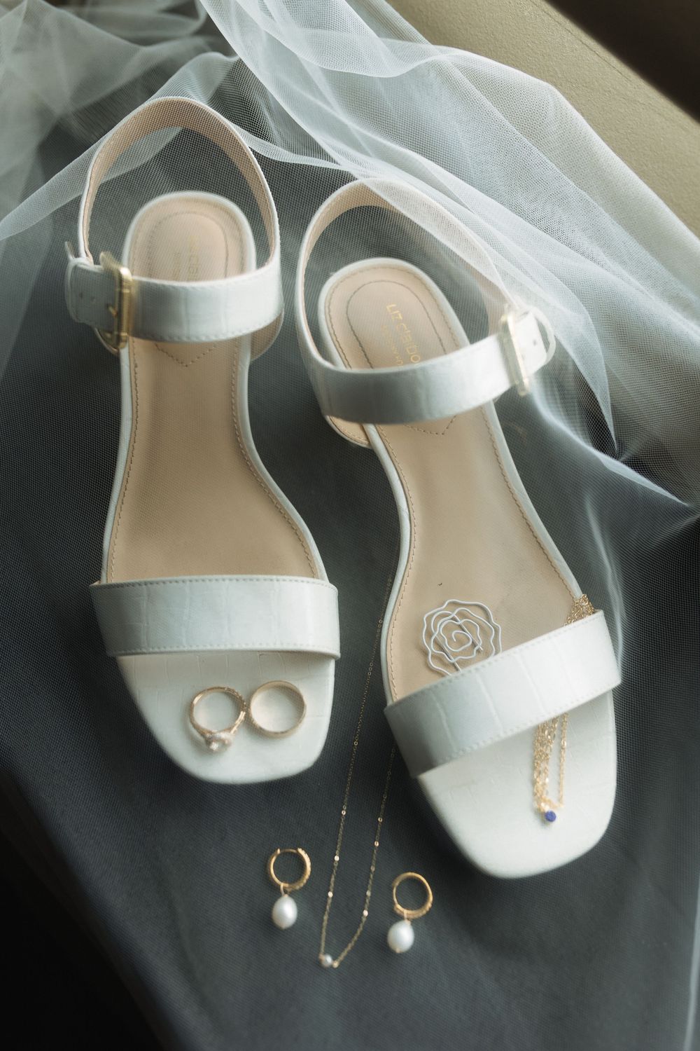 Elegant white bridal sandals displayed with wedding rings and pearl jewelry on flowing tulle fabric.
