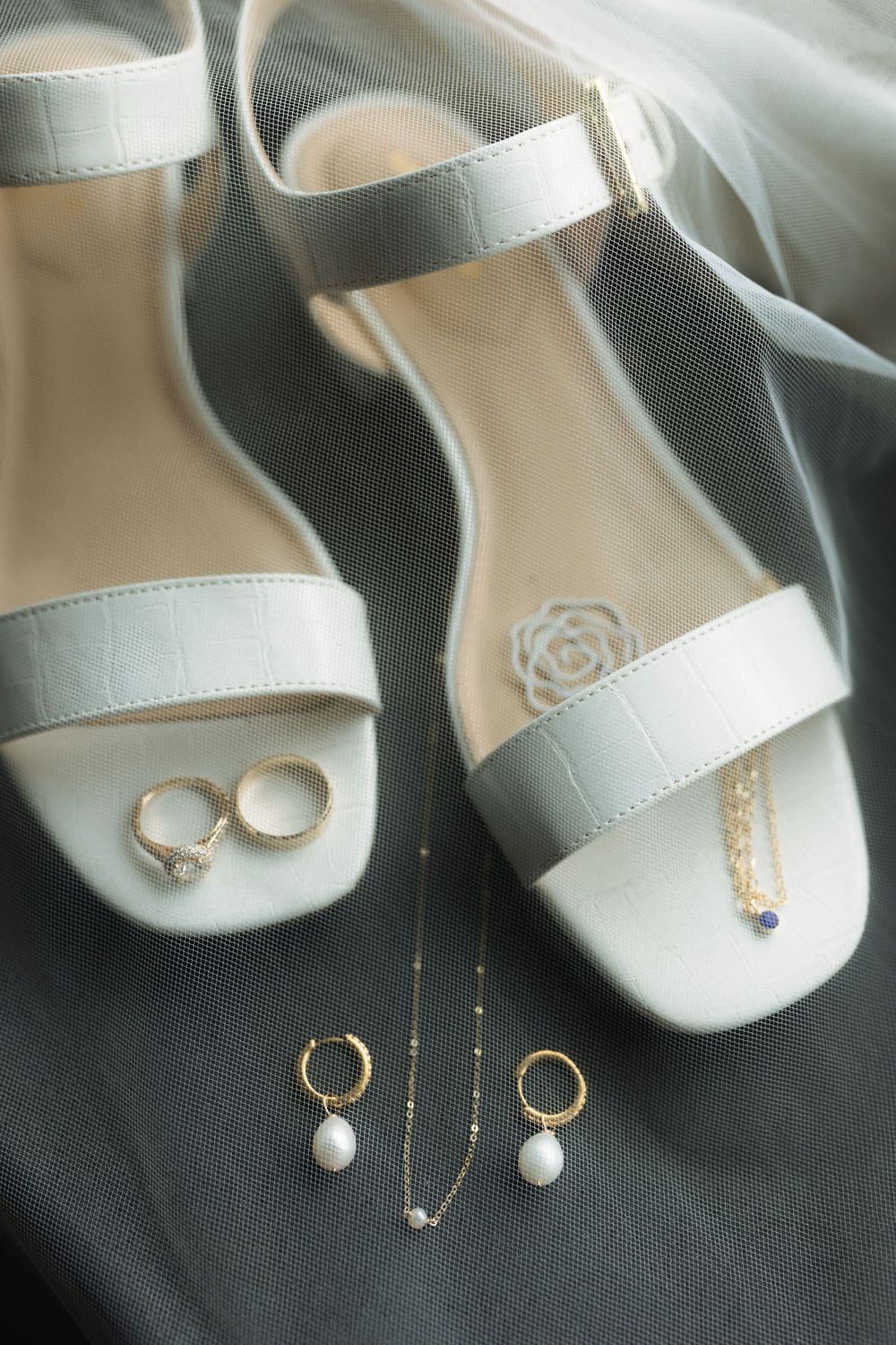 White bridal sandals with matching pearl and gold jewelry accessories arranged on dark fabric.