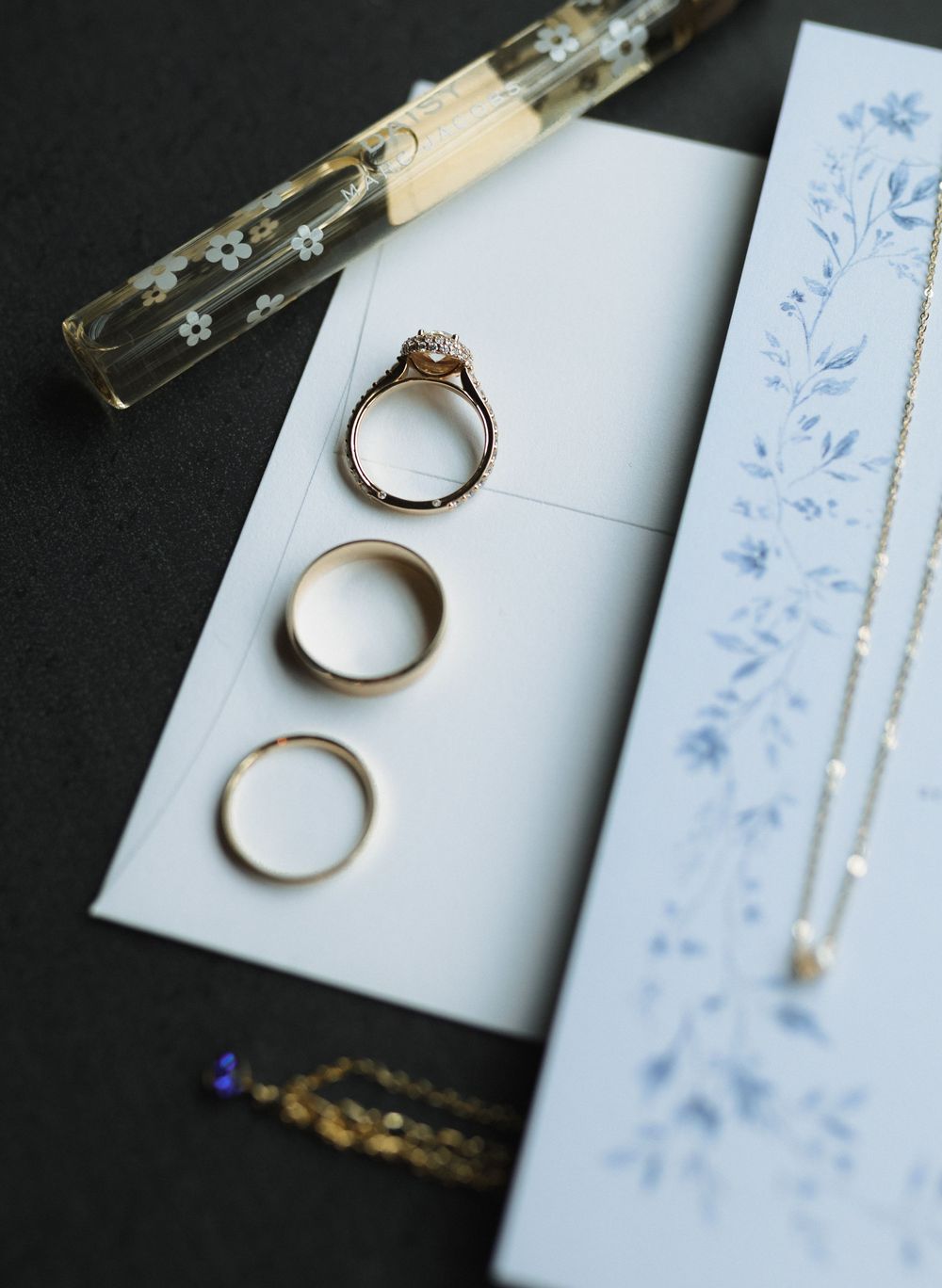 Three gold wedding rings arranged on white paper beside a delicate blue floral pattern and glass vial.