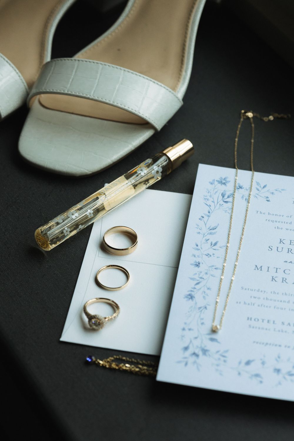 Wedding details including white bridal shoes, gold rings, perfume bottle and floral invitation on dark surface.
