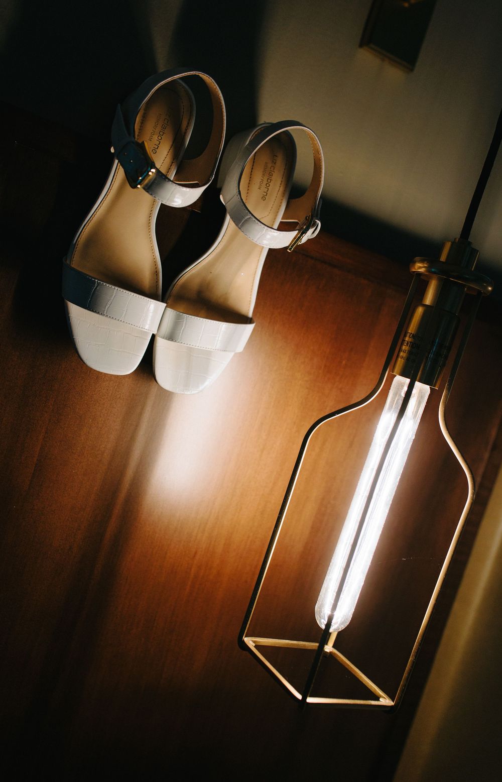 White leather sandals with ankle straps sit on a wooden surface next to a modern geometric light fixture.