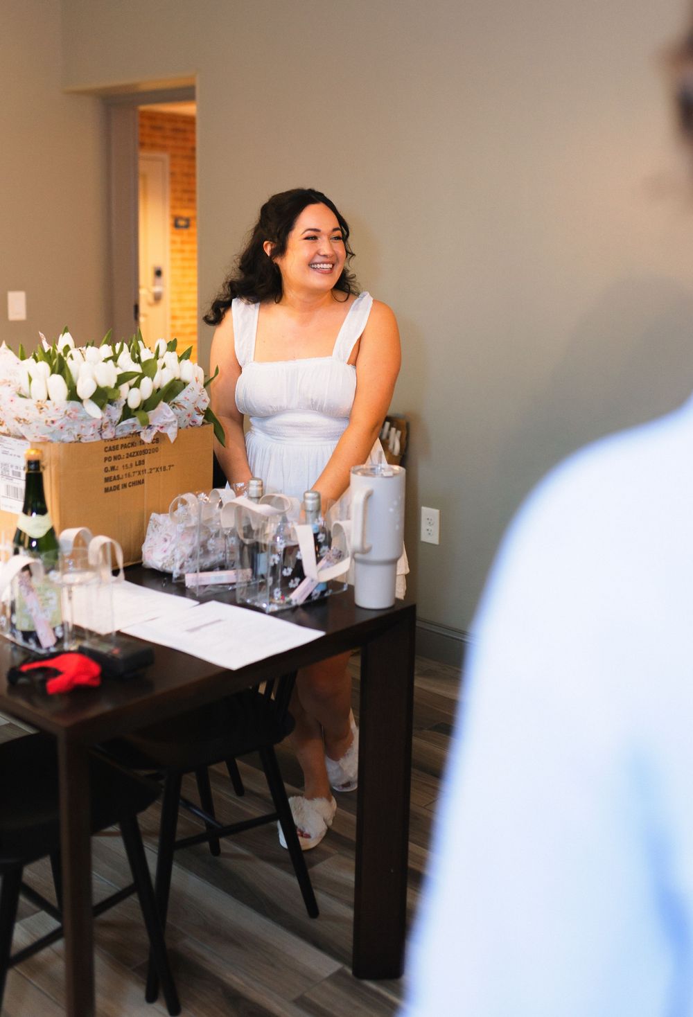 Person in a white dress stands smiling at a table with floral arrangements and party supplies.