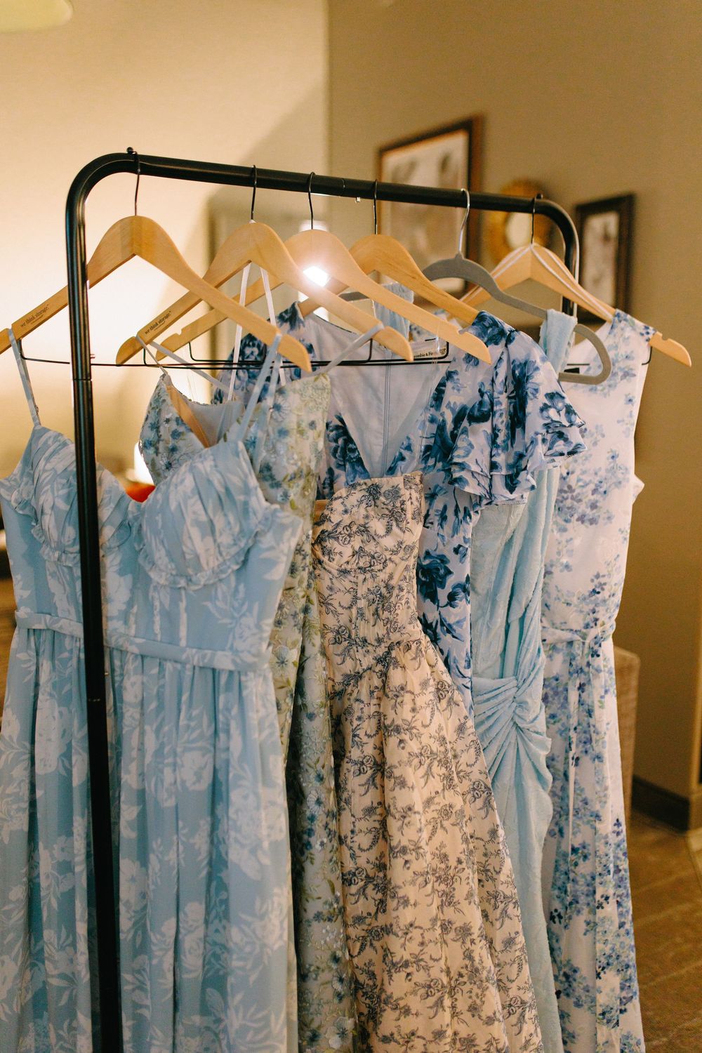 Row of light blue and floral print bridesmaids dresses hanging on wooden hangers against a beige wall.