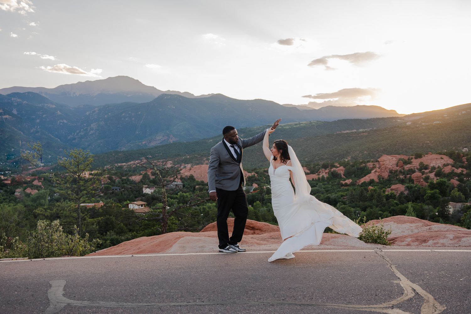Couple dances joyfully on red rocks with stunning mountain views and sunset sky behind them.