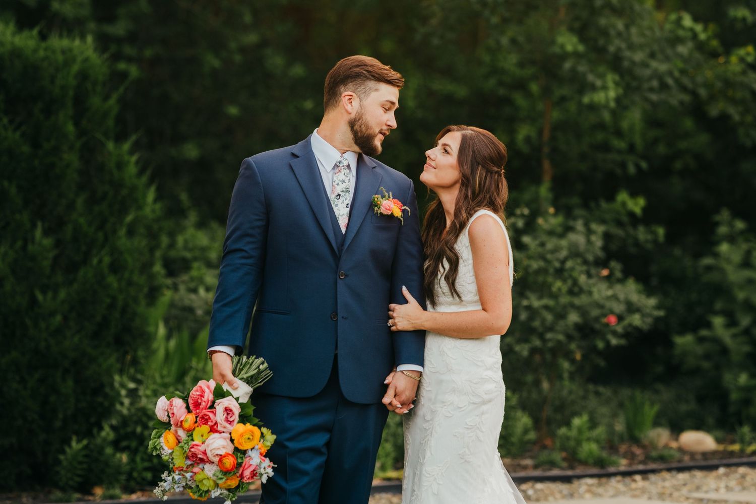 A loving couple in wedding attire share a tender moment outdoors while holding a vibrant bouquet of flowers.