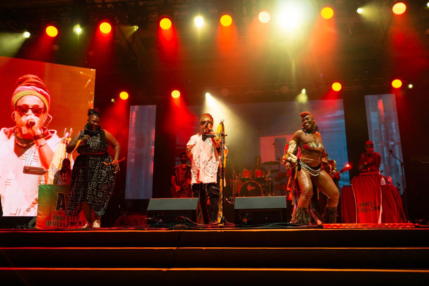Musical performers on an illuminated stage with red and yellow lighting performing in front of digital display screens.