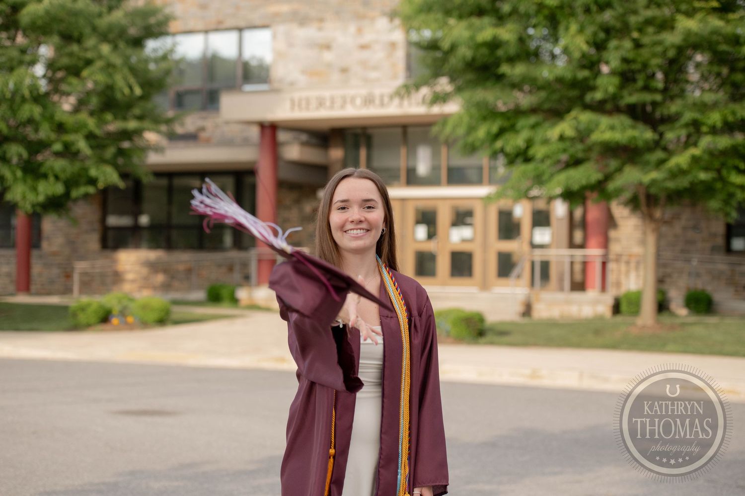 A graduate in maroon cap and gown celebrates graduation day in an outdoor campus setting.