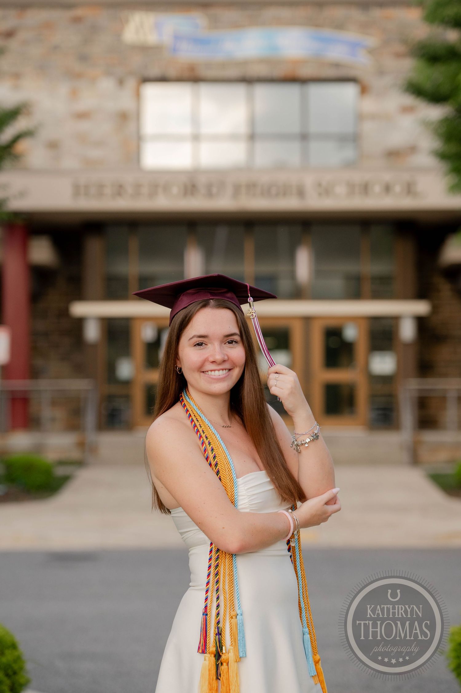 A graduate wearing white dress with gold honor cords stands outside a stone school building entrance.