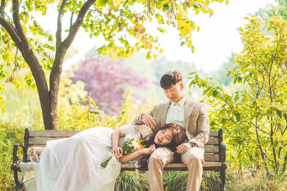 Couple in wedding attire resting on wooden bench in autumn garden setting.