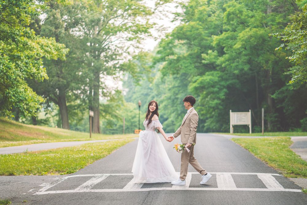 Couple in wedding attire holding hands while crossing street in green park setting.