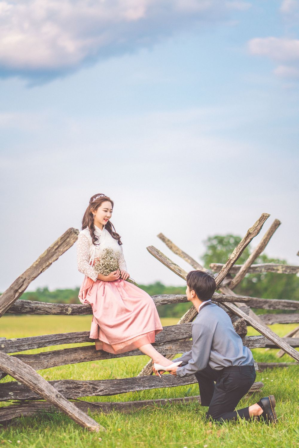 Couple in vintage attire pose romantically by rustic wooden fence in countryside setting.