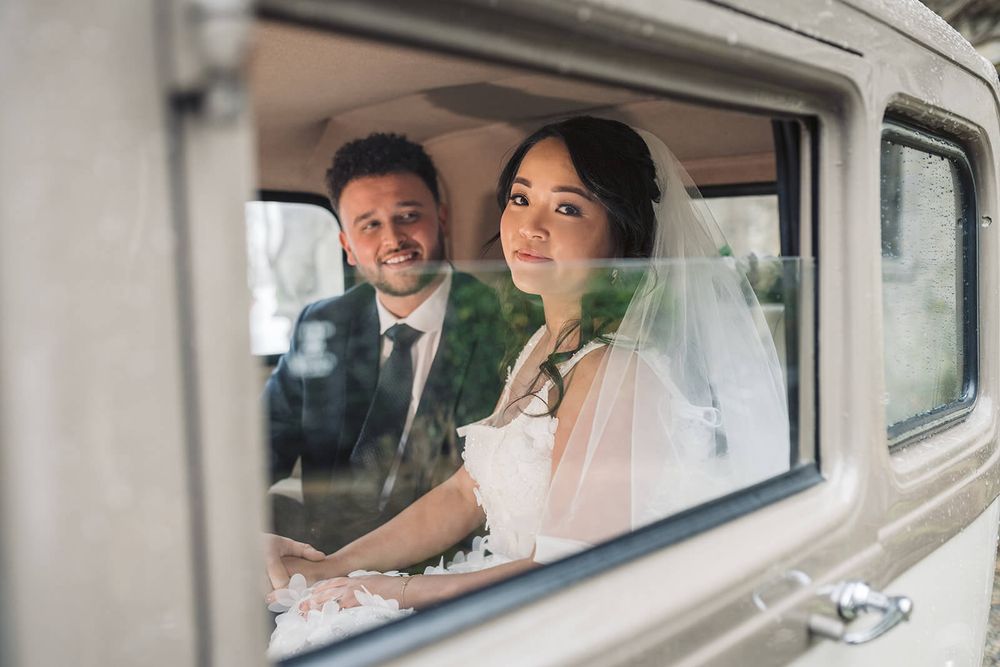Newlyweds sit together in a vintage car viewed through the window on their wedding day, captured by Dina Ozola Photography.