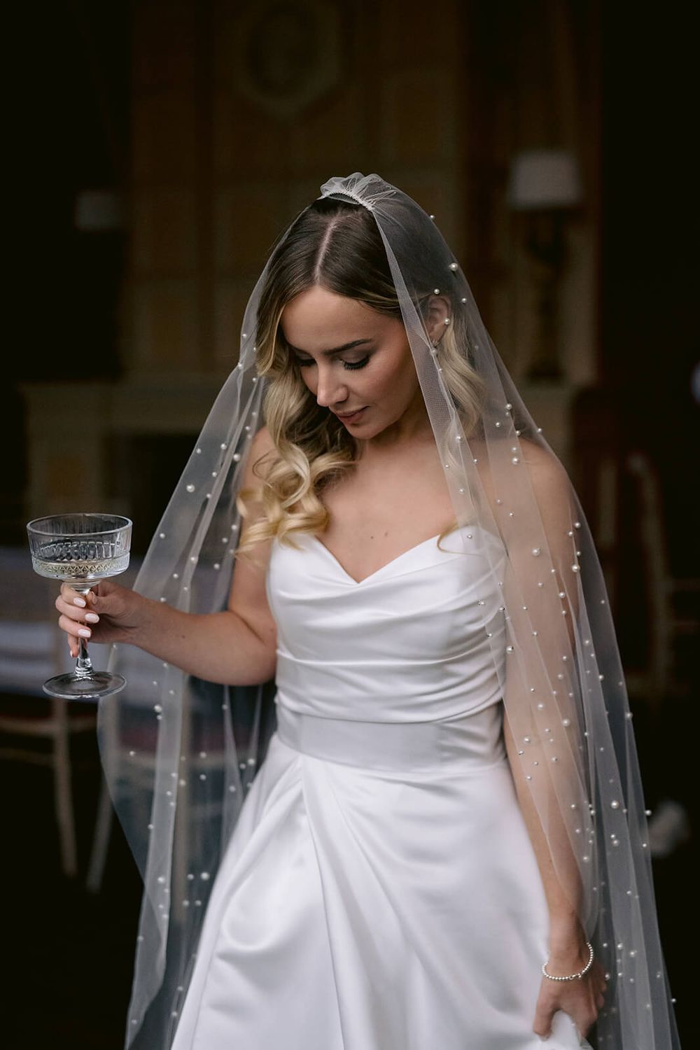 Bride in white strapless gown holds champagne glass while wearing a pearl-adorned veil, captured by Dina Ozola Photography.